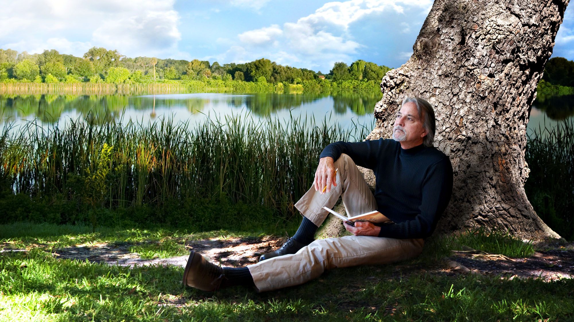 Man reading under a tree by a serene lake with lush greenery