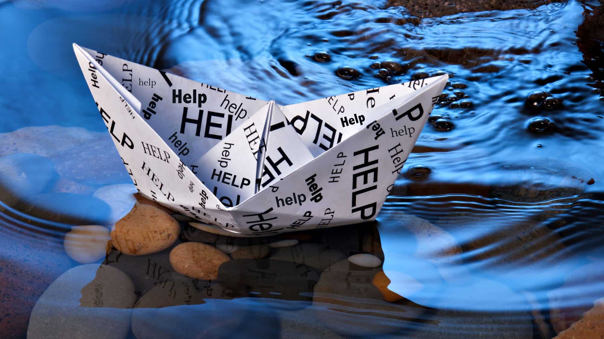 Paper boat with 'help' text floating on a stream surrounded by rocks