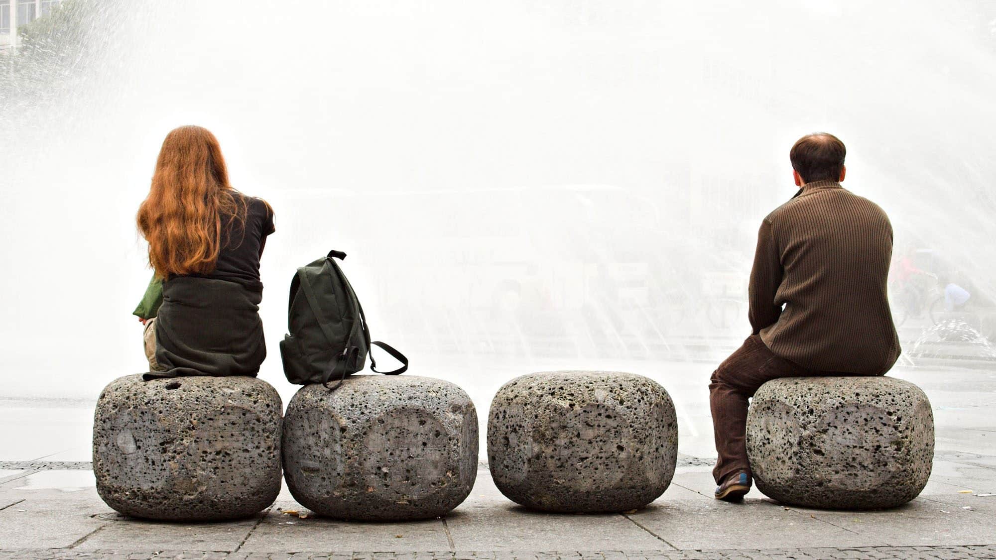 Two people sitting back to back on stone benches near a water fountain